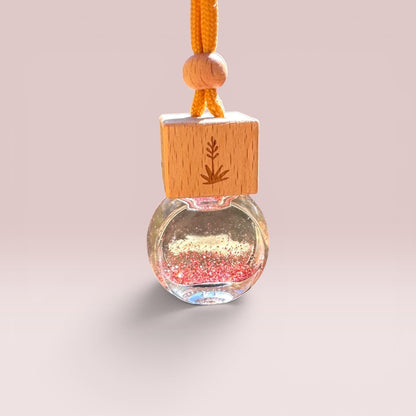 A small hanging ornament with a clear round glass bottle filled with glittery pink and gold liquid, topped with a light wood cube engraved with a leaf, and suspended by an orange cord with wooden beads.