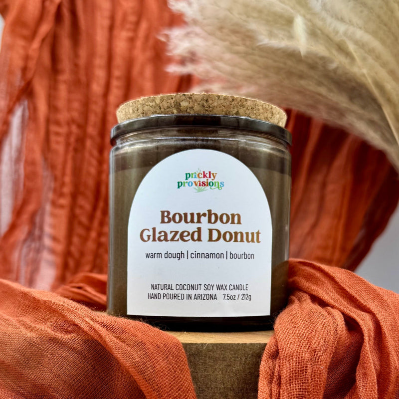 Candle jar labeled 'Bourbon Glazed Donut' with a cork lid, held by orange fabric.