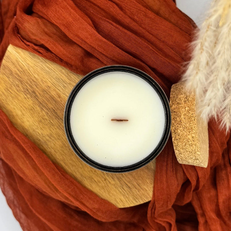 Candle in a black container on a wooden surface with orange fabric and a textured object.