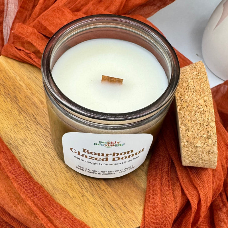 Candle in a glass jar with a wooden wick, surrounded by orange fabric on a wooden surface.