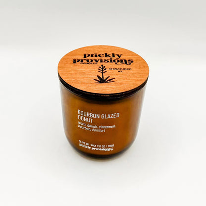 A brown glass jar candle labeled 'Bourbon Glazed Donut' with white wax, indicating a scent of warm dough, cinnamon, bourbon, and comfort.