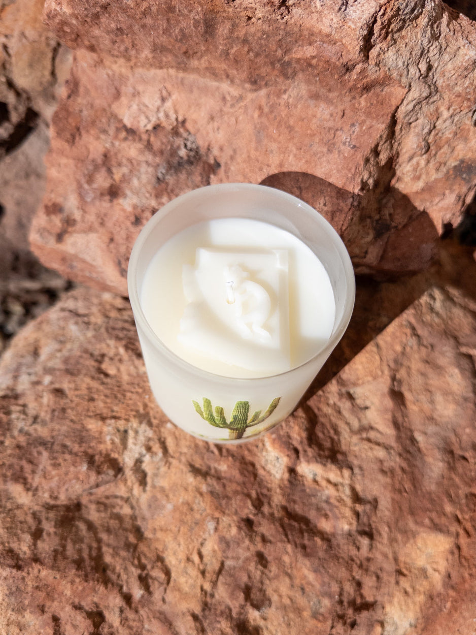 Candle in a white container with a green saguaro cactus design on a textured brown surface