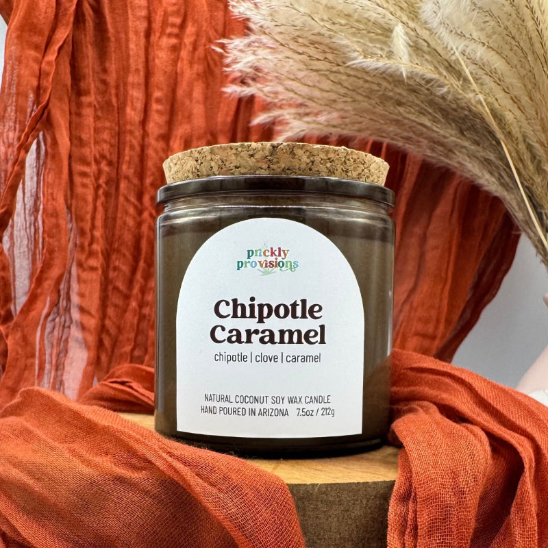 Candle jar labeled 'Chipotle Caramel' with a cork lid on an orange fabric background.