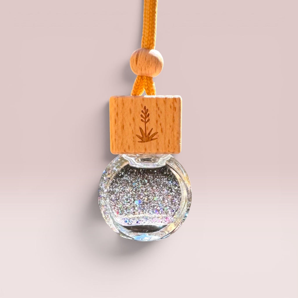 A glass car air freshener bottle with a clear round bottom chamber filled with silver holographic glitter, topped with a square light wood block engraved with a plant design, and suspended by an orange cord on a soft beige background.