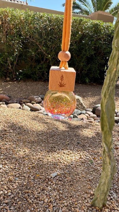 A hanging glass diffuser bottle with a wooden top block and rope loop, placed outdoors on a gravel surface with hedges and cacti in the background.