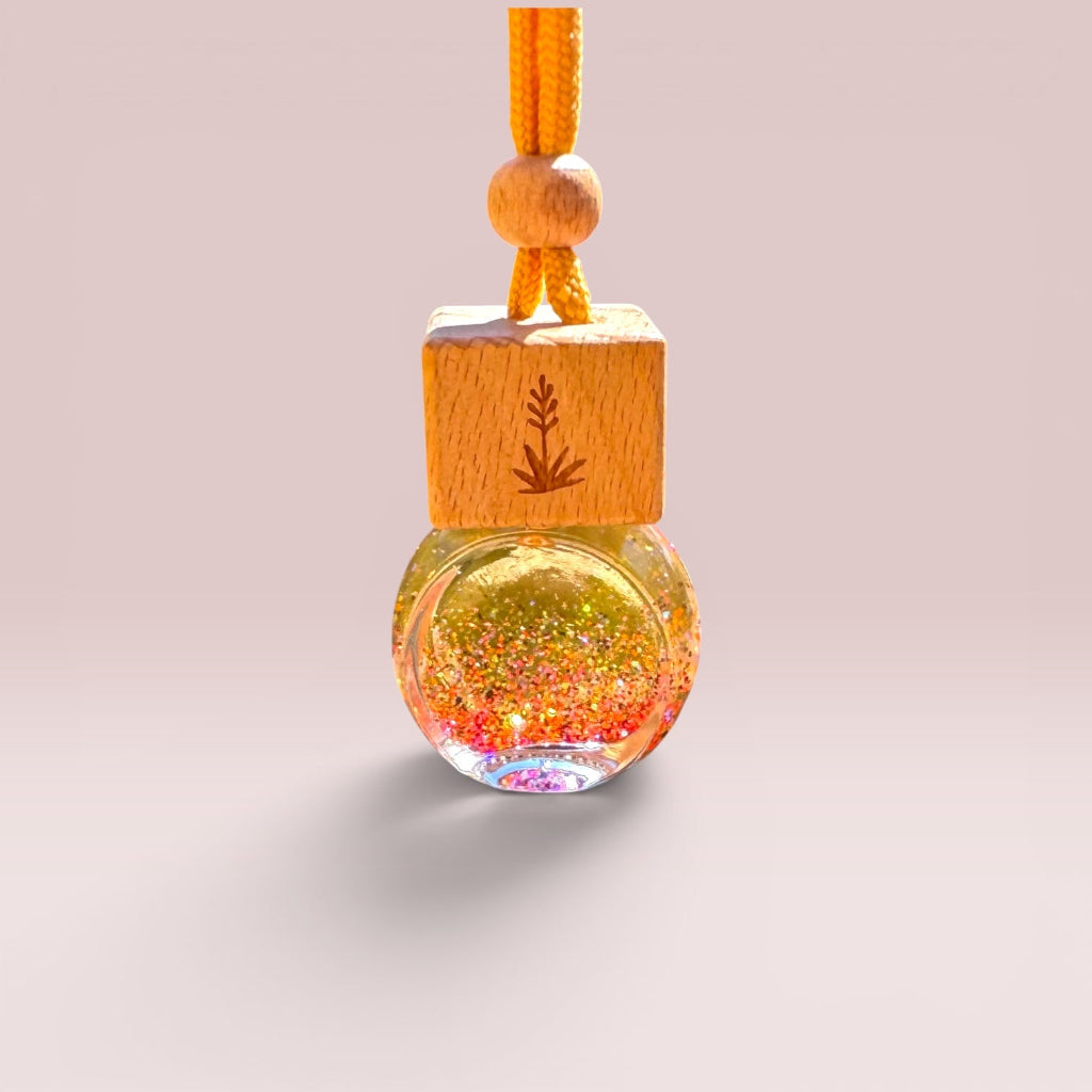A hanging glass bottle ornament with a wooden cap and orange cord, filled with glittering gold, pink, and iridescent liquid, shown on a soft beige background.