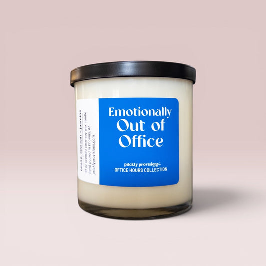 A jar candle labeled 'Emotionally Out of Office' by prickly provisions, placed on a dark wooden desk with a blurred laptop in the background.