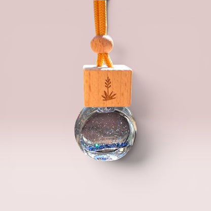 A glass bottle pendant with a wooden top block engraved with a pine tree, filled with glittery clear liquid and blue beads, hanging from an orange cord on a light beige background.