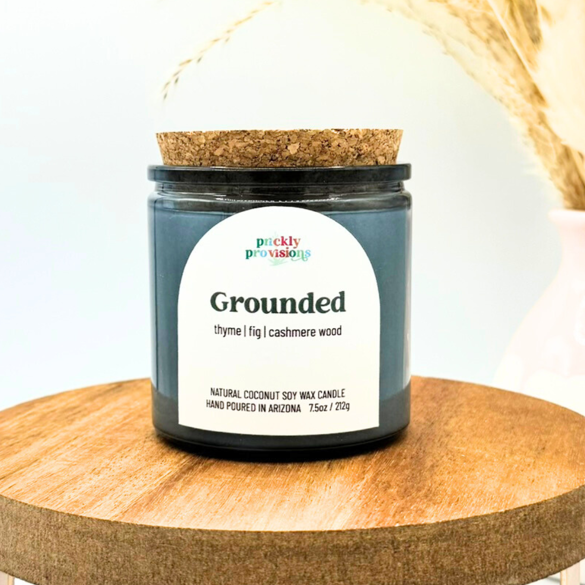 Candle jar labeled 'Grounded' by Prickly Provisions on a wooden surface with a blurred background.