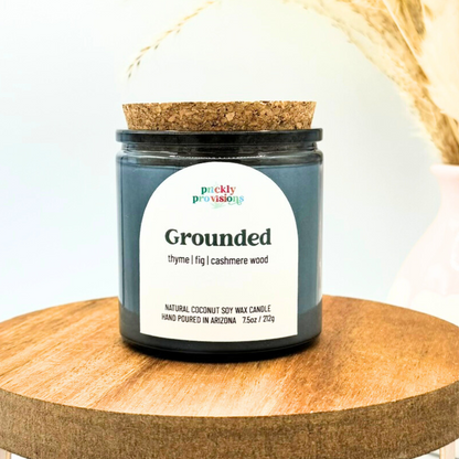 Candle jar labeled 'Grounded' by Prickly Provisions on a wooden surface with a blurred background.