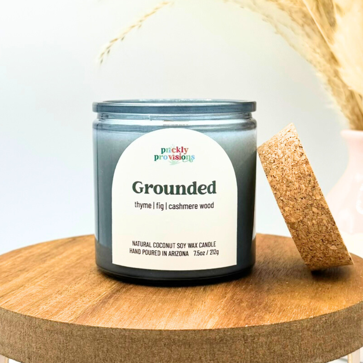 Candle labeled 'Grounded' by Prickly Provisions on a wooden surface with a blurred background