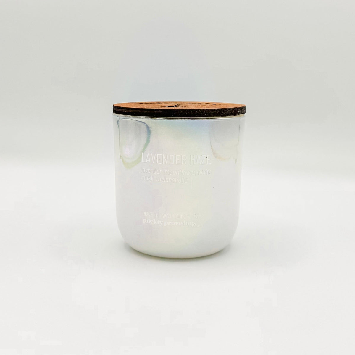 An opal white iridesent glass candle labeled 'Lavender Haze' with additional text indicating olive oil, lavender, moonflower, white musk, melancholia. The candle is in a glass jar with a wooden lid.