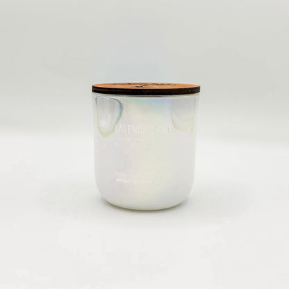 An opal white iridesent glass candle labeled 'Lavender Haze' with additional text indicating olive oil, lavender, moonflower, white musk, melancholia. The candle is in a glass jar with a wooden lid.