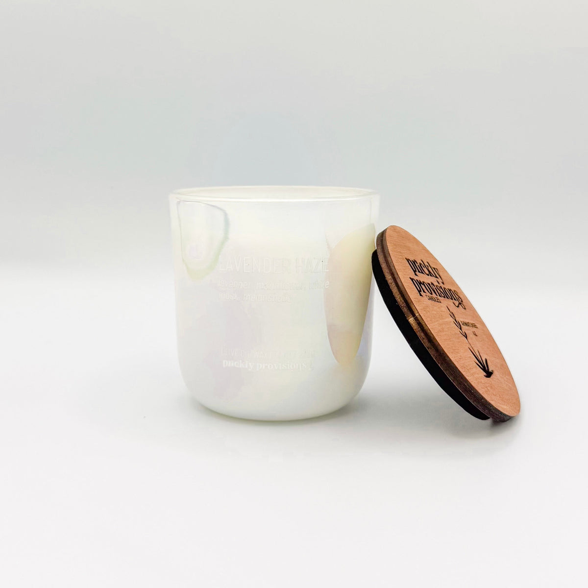 An opal white iridesent glass candle labeled 'Lavender Haze' with additional text indicating olive oil, lavender, moonflower, white musk, melancholia. The candle is in a glass jar with a wooden lid.
