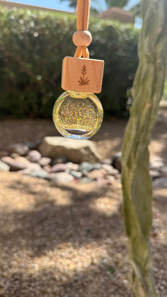 A small hanging ornament or pendant with a round clear bubble filled with gold glitter, attached to a tan cord and a square wooden tag engraved with a plant symbol, photographed outdoors against a blurred garden background.