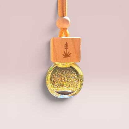 A small amber glass bottle filled with sparkling gold glitter, attached to a light wood block engraved with a leaf design and a matching wooden bead, hanging from an orange cord against a soft beige background.