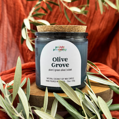 Candle jar labeled 'Olive Grove' by 'prickly provisions' with olive leaves around on a warm background.