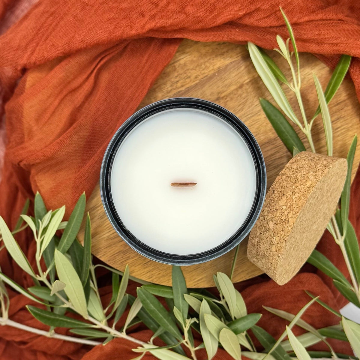 White candle in a glass container on a wooden board with green olive leaves and orange fabric