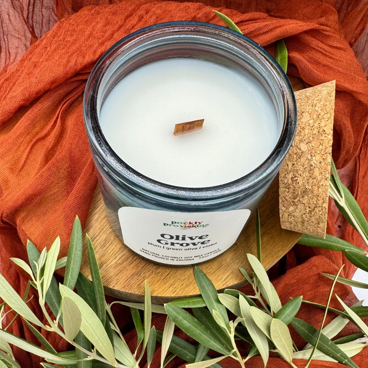 Candle in a glass jar with a wooden wick, surrounded by green olive tree leaves and orange fabric.