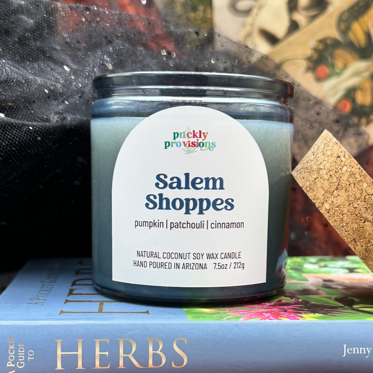 Candle labeled 'Salem Shoppes' by Prickly Provisions on a book with a blurred background