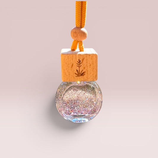 A hanging car air freshener or perfume bottle with a round clear glass body filled with iridescent glitter, a light wood cube with a small leaf engraving, and an orange cord at the top, shown on a soft beige background.