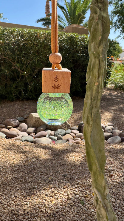 A hanging glass ornament or diffuser with a light wood cap engraved with a leaf design, suspended outdoors in front of a cactus and garden background.
