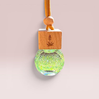 A hanging ornament or pendant with a round clear glass globe filled with iridescent green glitter, topped by a square light wood block engraved with a leaf, and suspended from an orange cord on a soft pink background.