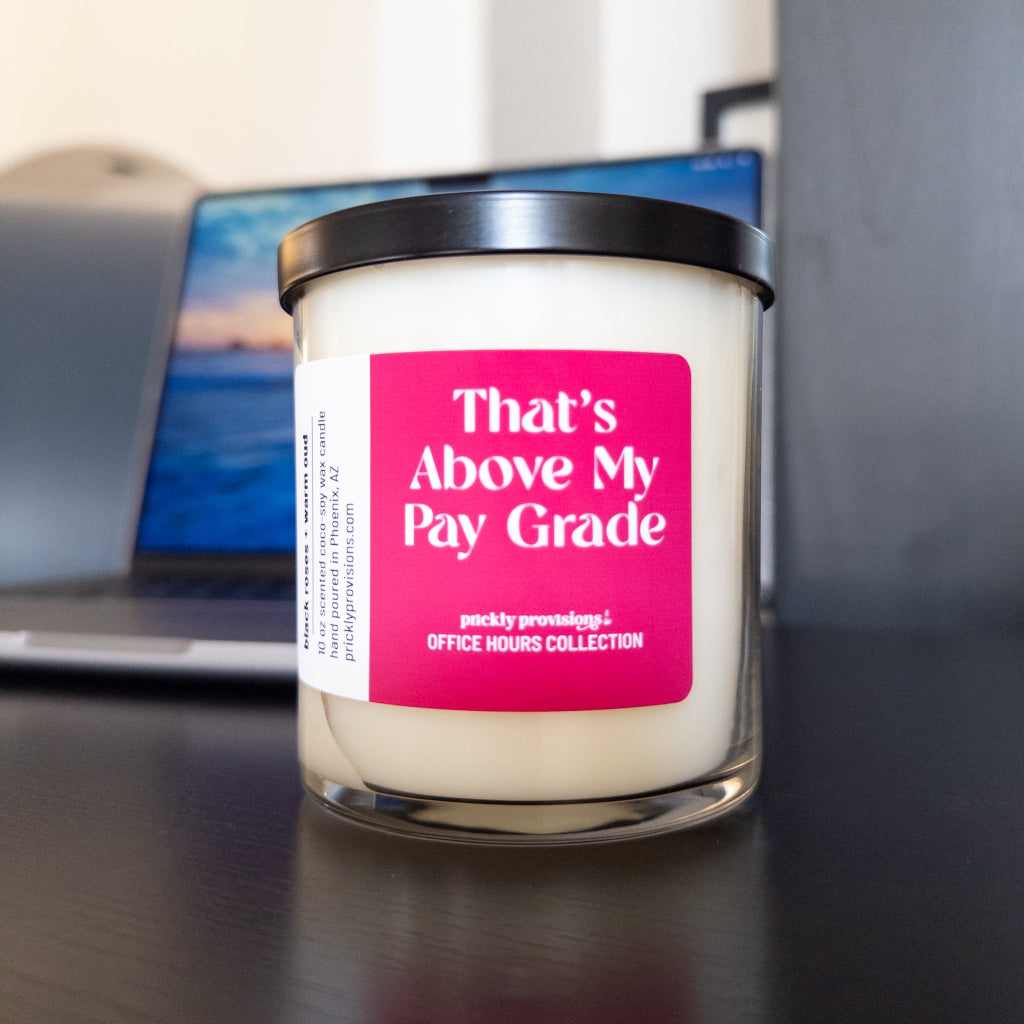 A jar candle with cream-colored wax and a black lid sits on a dark desk in front of a blurred laptop. The candle features a bright pink label with white text that reads 'That's Above My Pay Grade' and 'prickly providence: OFFICE HOURS COLLECTION'.
