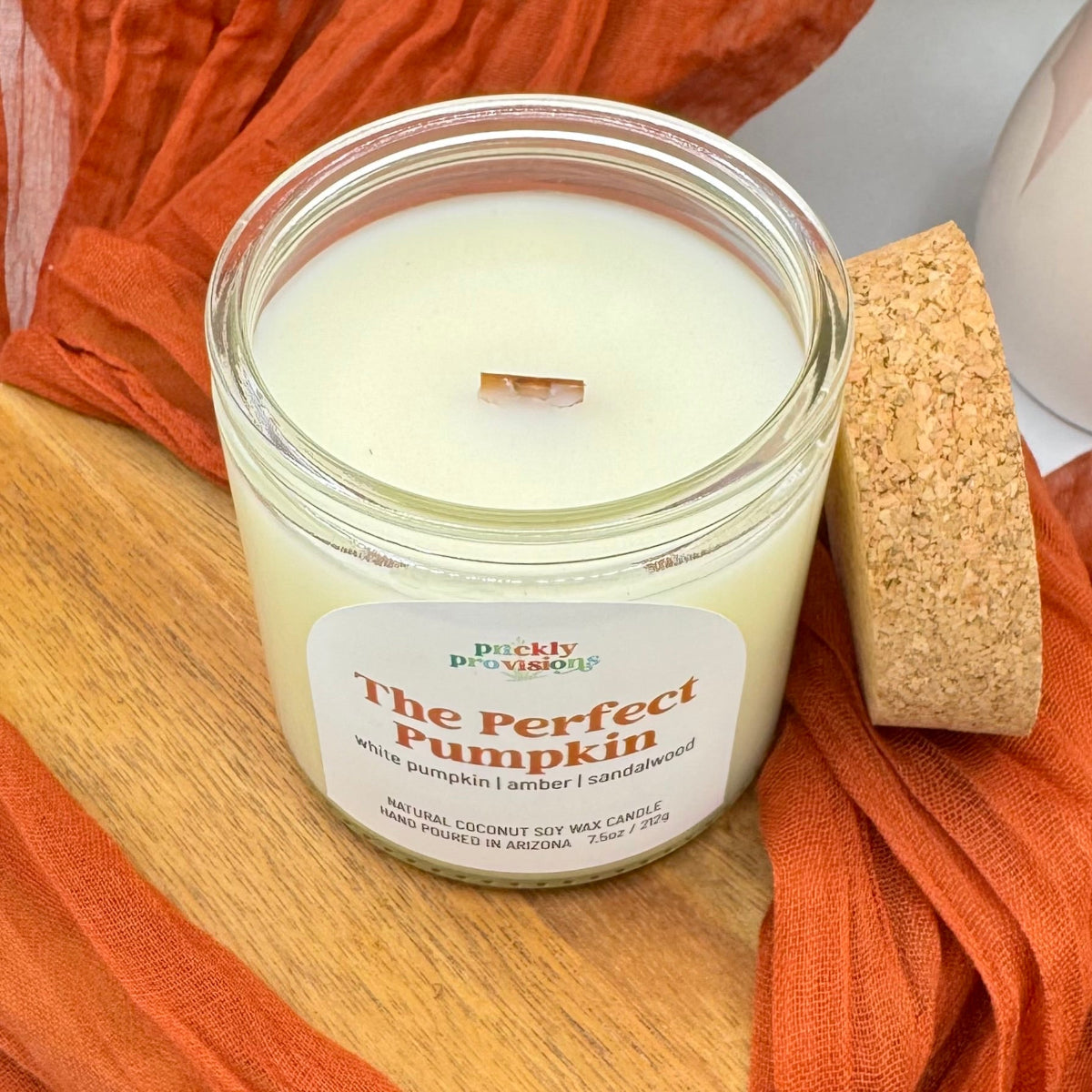 Candle labeled 'The Perfect Pumpkin' by Prickly Provisions on a wooden surface with orange fabric.