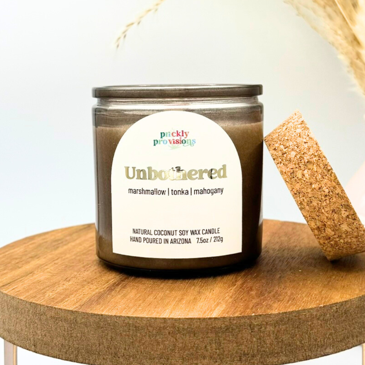 Candle jar labeled 'Unbothered' on a wooden surface with a cork coaster.