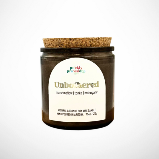 Candle jar with cork lid and 'Unbothered' label on a white background