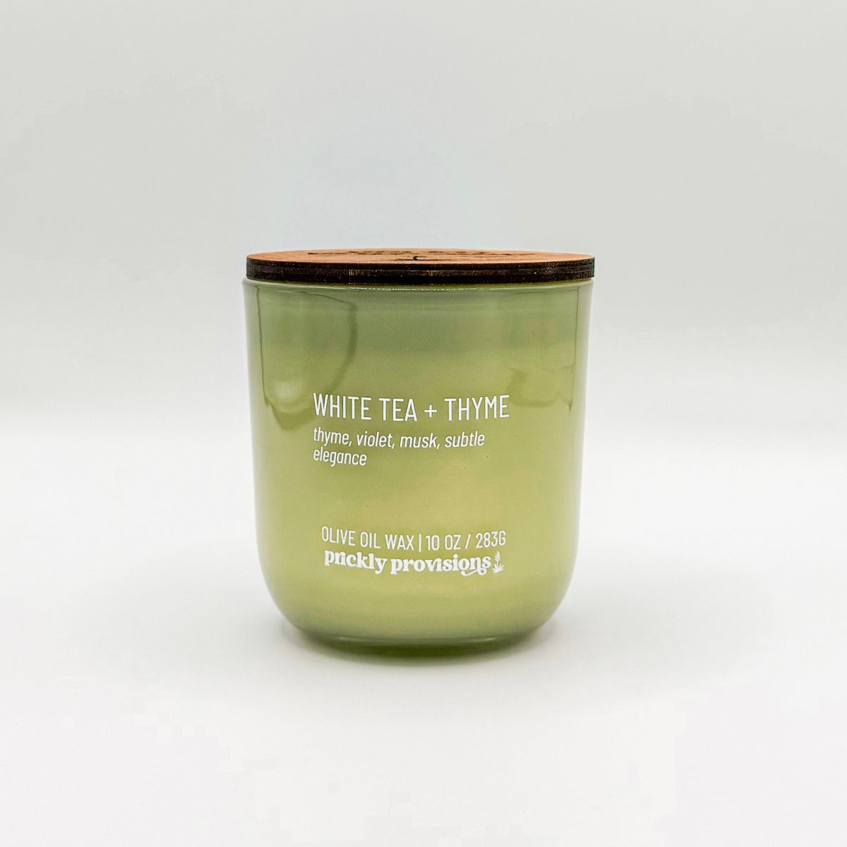 A sage green glass candle labeled 'White Tea and Thyme' with additional text indicating olive oil, thyme, violet, musk, and subtle elegance. The candle is in a glass jar with a wooden lid.