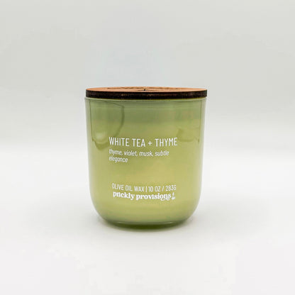 A sage green glass candle labeled 'White Tea and Thyme' with additional text indicating olive oil, thyme, violet, musk, and subtle elegance. The candle is in a glass jar with a wooden lid.