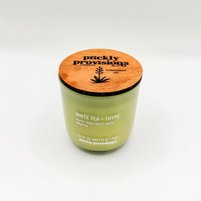 A sage green glass candle labeled 'White Tea and Thyme' with additional text indicating olive oil, thyme, violet, musk, and subtle elegance. The candle is in a glass jar with a wooden lid.