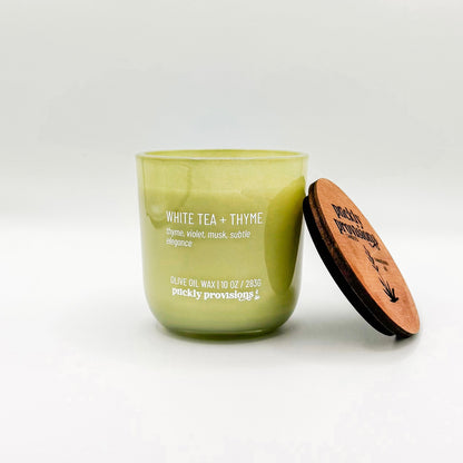 A sage green glass candle labeled 'White Tea and Thyme' with additional text indicating olive oil, thyme, violet, musk, and subtle elegance. The candle is in a glass jar with a wooden lid.