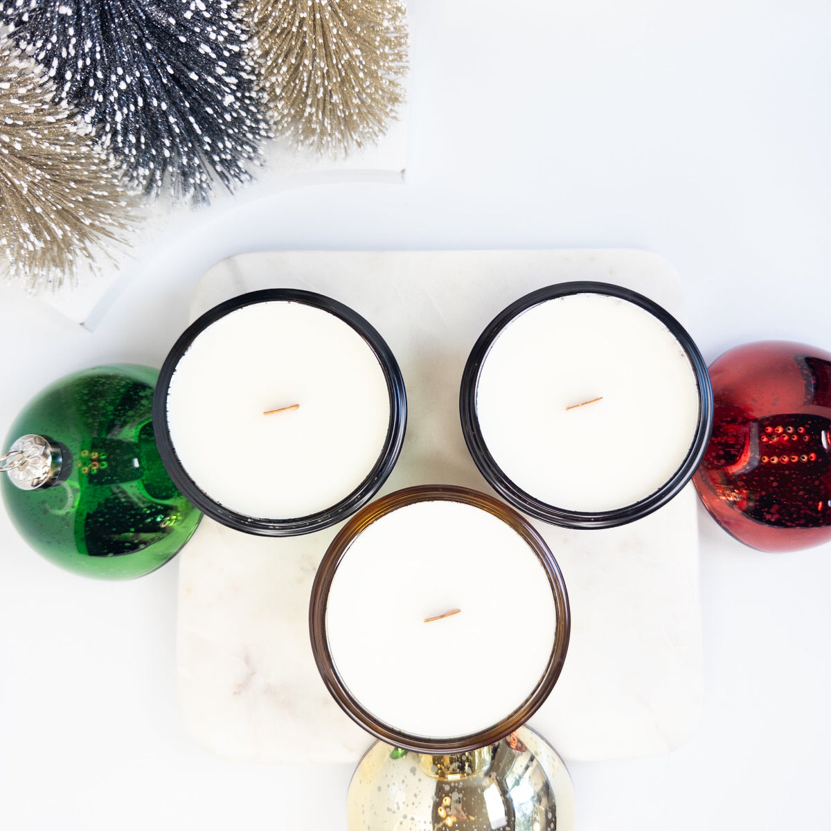 Three Christmas ornament candles with wood wicks on a white surface