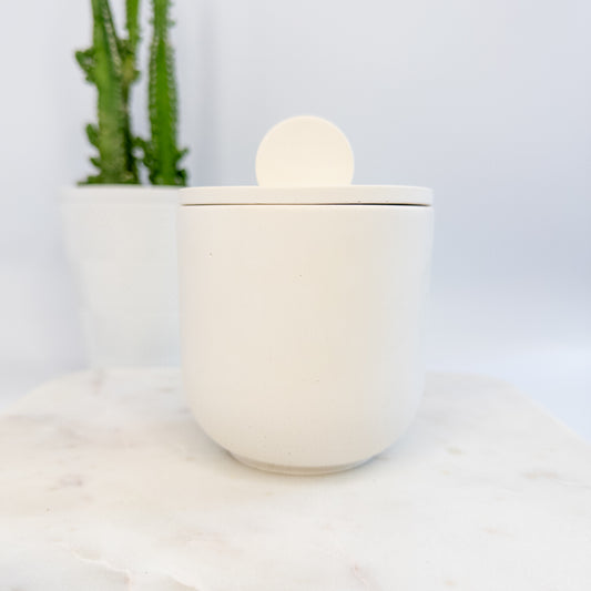 White cement candle  jar with a lid on a white surface with a blurred plant in the background