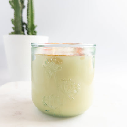 Glass candle with floral design on a white surface with a plant in the background