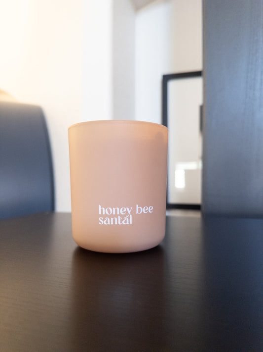 Khaki candle labeled 'honey bee santal' on a dark surface with a blurred background