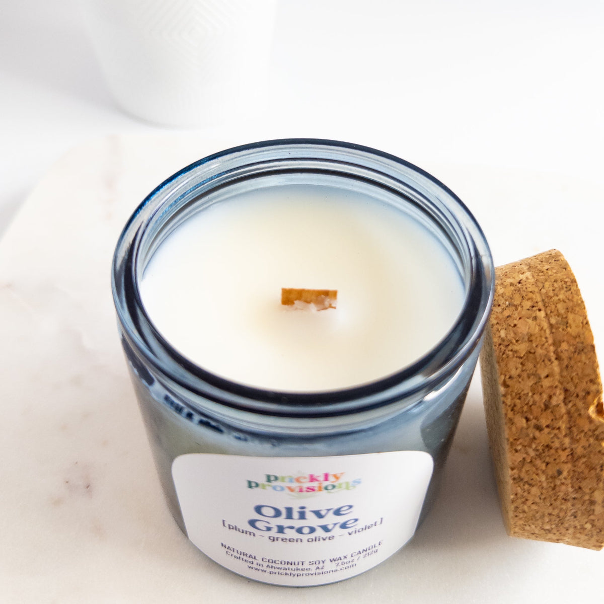 Candle in a glass jar labeled 'Olive Grove' with a cork lid on a white background