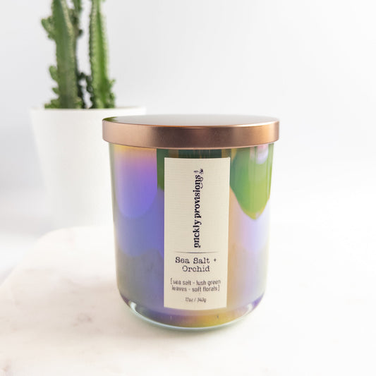 Candle with a colorful label and rose gold lid on a white surface with a plant in the background